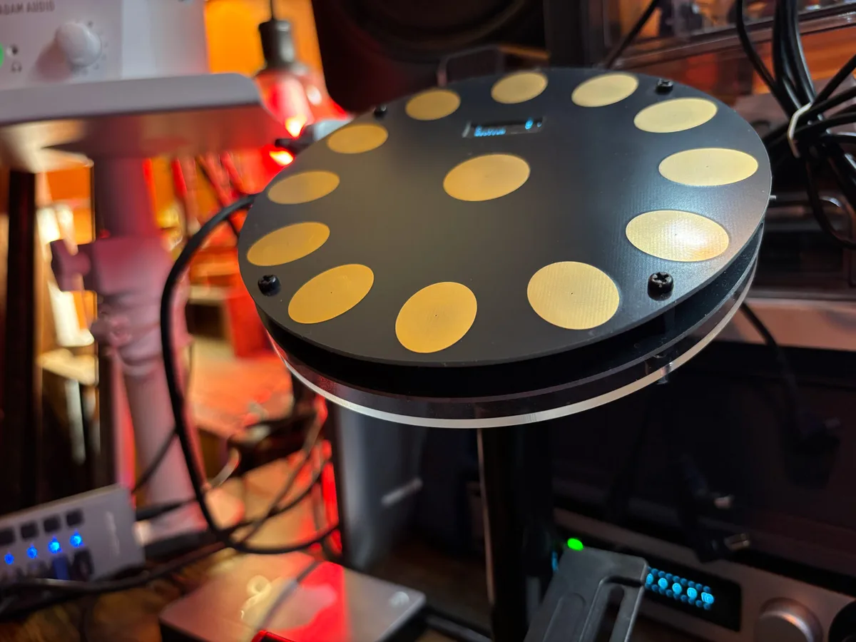 A round electronic MIDI percussion controller with twelve tan pads arranged around a small display, mounted on a stand in a studio.