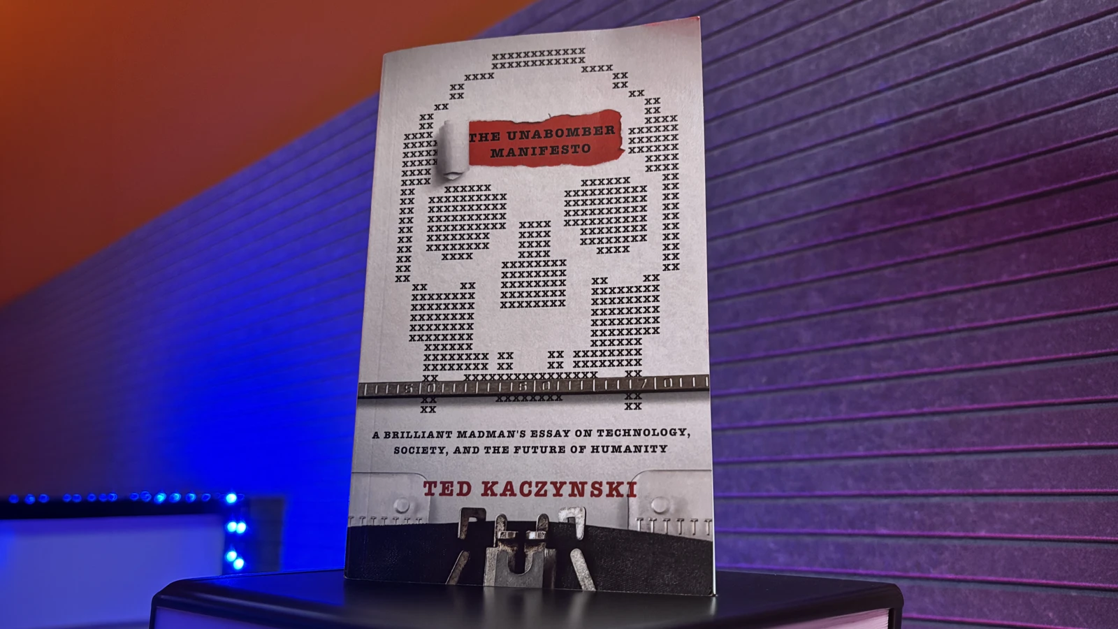 A paperback copy of “The Unabomber Manifesto” by Ted Kaczynski, standing upright against an orange-and-purple backdrop.